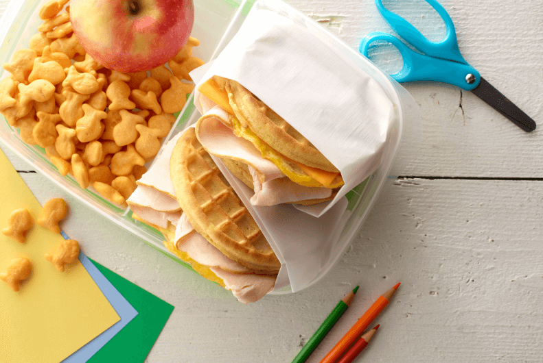 Turkey waffle sandwich wrapped on a tray with goldfish and an apple 